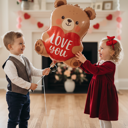 Two children holding a large teddy bear-shaped balloon with 'Love You' text in a home setting.